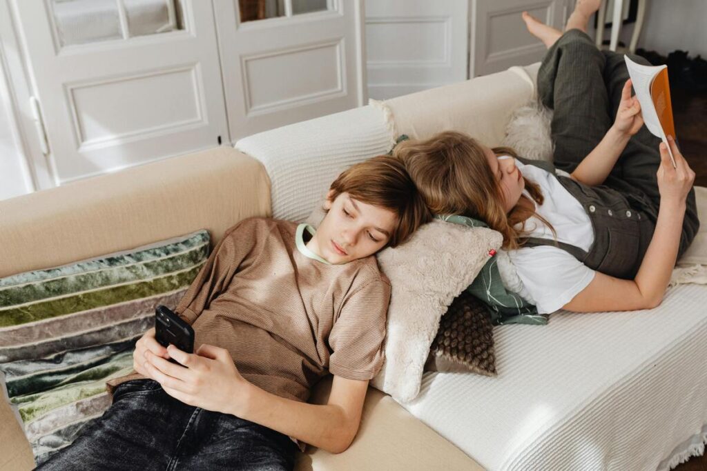 Dječak na kauču gleda sadržaj na mobitelu, a djevojčica na kauču čita knjigu