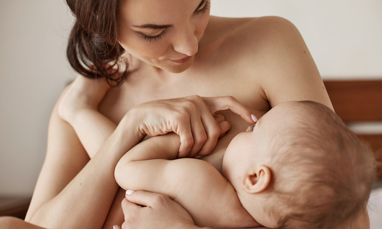 Young tender nude mother breastfeeding hugging her newborn baby sitting in bed at morning.